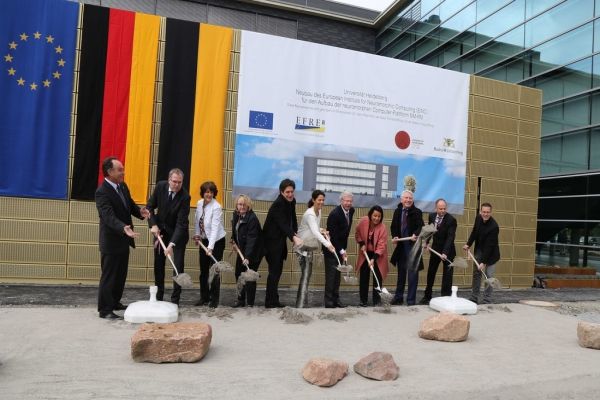 With German and EU flags in the background, representatives from the sponsoring authorities are using shovels to throw soil to the ground where the building is going to be constructed.